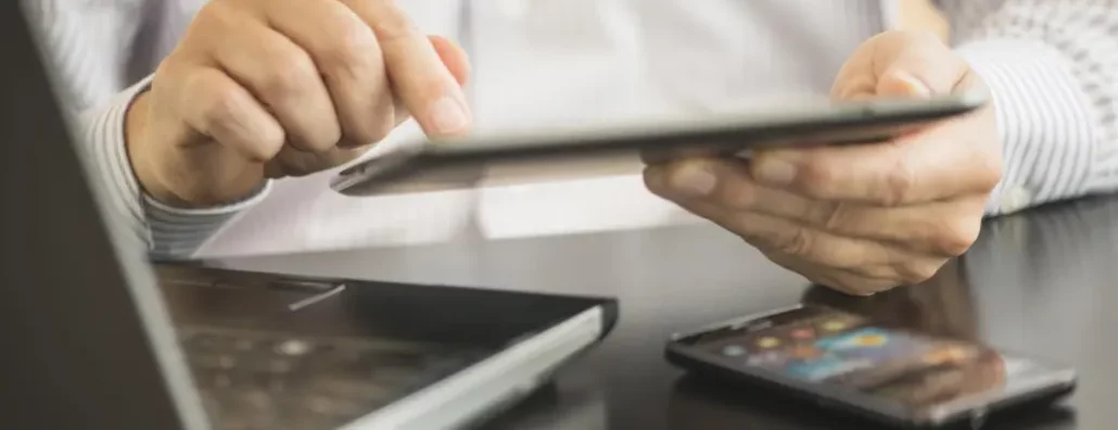 Manos de una persona utilizando una tablet, con un portátil y un smartphone sobre la mesa.