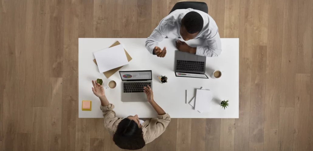 Vista cenital de dos personas trabajando en una mesa blanca, con laptops, hojas de papel, café y una planta pequeña, sobre un suelo de madera.