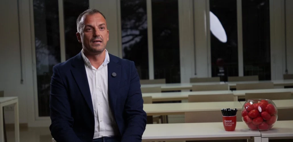 Xavier Rodríguez assegut en una aula moderna amb taules i cadires blanques, acompanyada d'un recipient amb pilotes vermelles i un got amb bolígrafs sobre una taula.