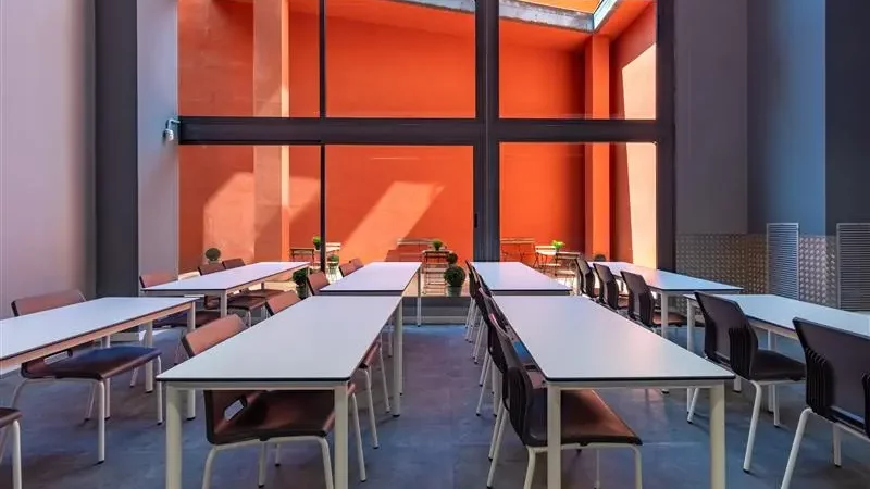 Aula moderna con mesas y sillas, grandes ventanales que dan a un patio exterior con paredes de color naranja.