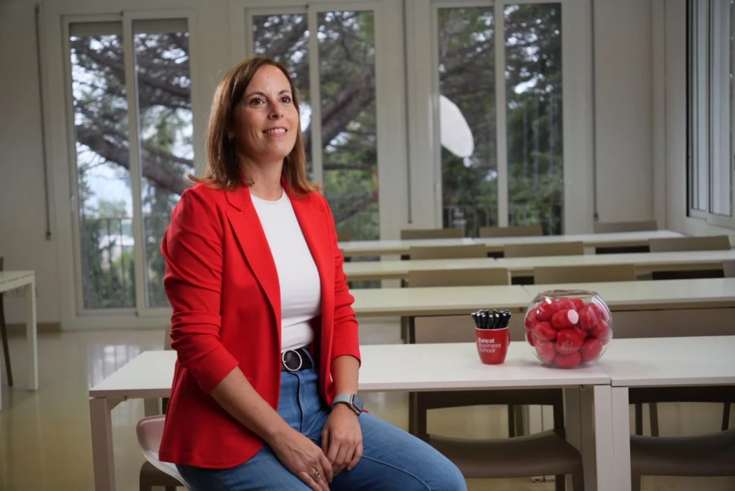 Mujer sentada en un aula luminosa, con una chaqueta roja, camiseta blanca y jeans, junto a un recipiente con bolas rojas y un vaso con bolígrafos sobre la mesa.