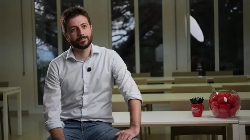 Hombre joven sentado en un aula moderna, con camisa blanca y jeans, en un ambiente educativo profesional.