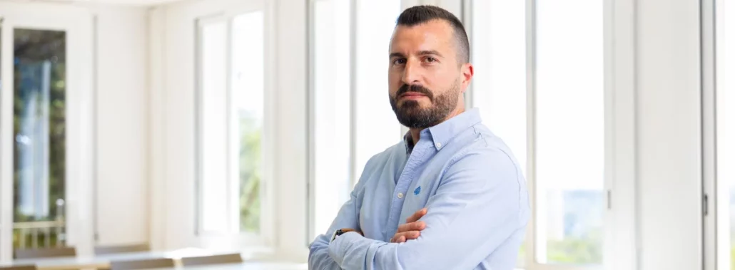 Hombre con camisa azul claro, de pie con los brazos cruzados, en un entorno profesional con ventanas grandes al fondo.