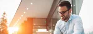 Hombre joven con gafas y camisa azul claro, sonriente mientras trabaja en un portátil al aire libre en un entorno moderno y luminoso.