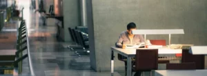 Estudiante sentado en una mesa iluminada mientras estudia en un espacio amplio y moderno.