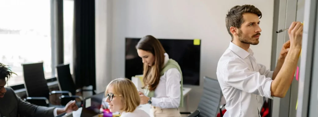 Grupo de personas trabajando en equipo en una oficina moderna, uno escribiendo en un tablero y otros revisando documentos