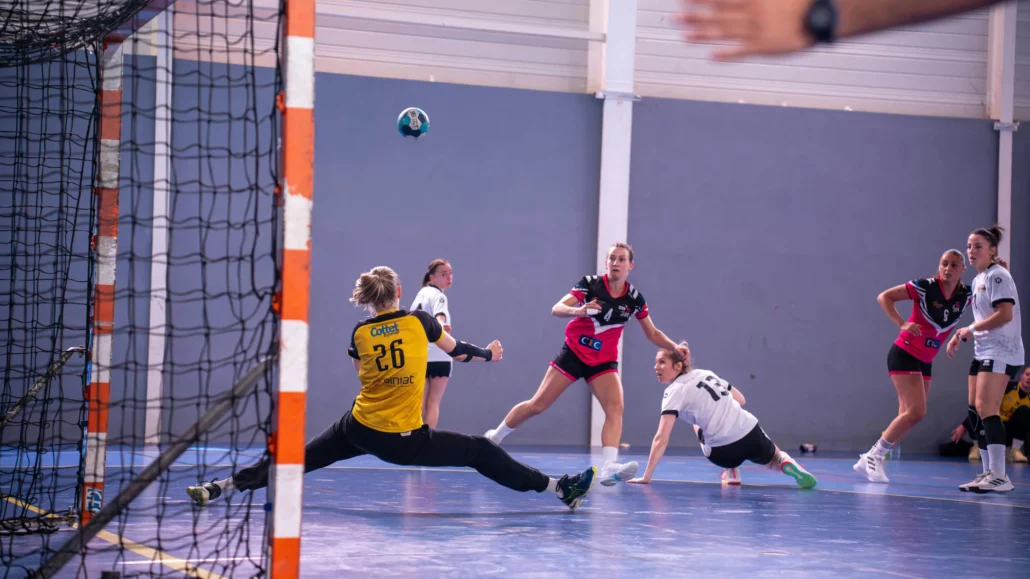 Partit d'handbol femení en acció, amb una jugadora llançant a porteria mentre la portera intenta aturar la pilota.