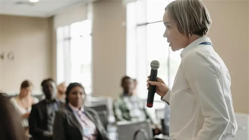 Mujer con cabello corto sosteniendo un micrófono, hablando frente a una audiencia en un entorno profesional o de conferencia.