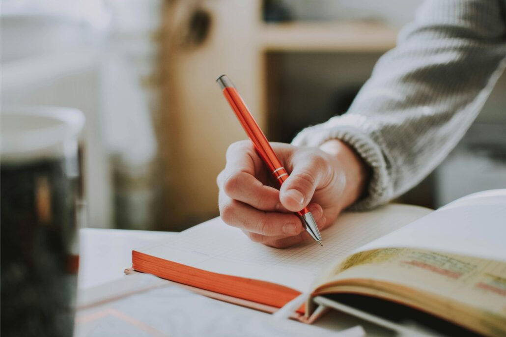 Mano de una persona escribiendo con bolígrafo rojo en un cuaderno de tapa naranja, sobre una mesa con libros y una taza