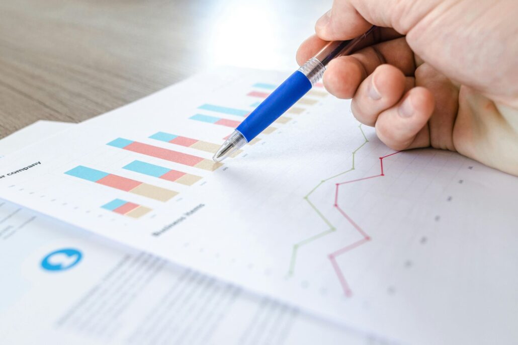 Mano sosteniendo un bolígrafo azul señalando gráficos impresos con datos financieros, incluyendo barras de colores y líneas de tendencia