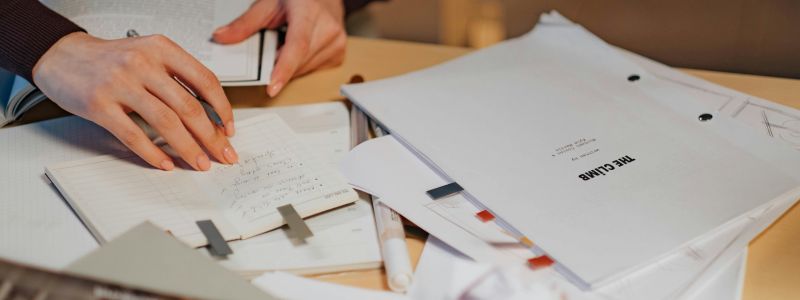 Persona revisando notas escritas a mano sobre una mesa llena de documentos, carpetas y material de oficina