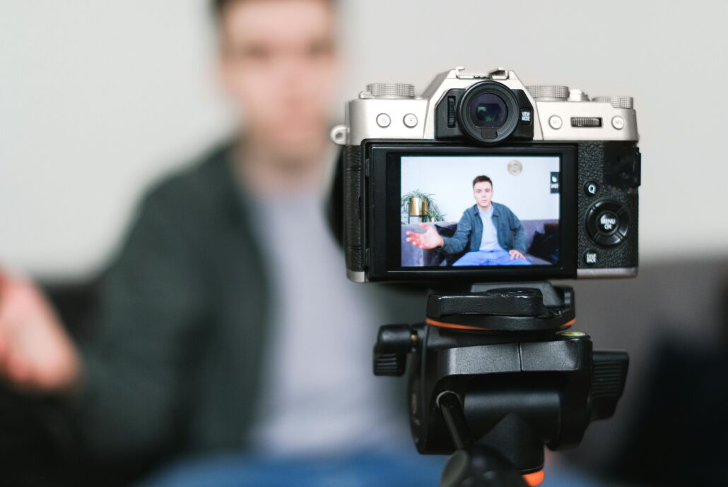 Persona grabando un video con una cámara sobre trípode, enfocada hacia él mientras está sentado hablando en un sofá